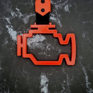 May include: An orange, 3D-printed engine symbol, commonly used as a car dashboard warning light. The symbol is attached to a black strap with two black screws. The background is a dark, marbled surface.