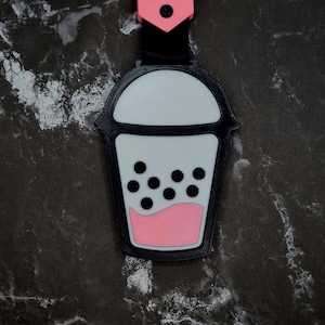 May include: A black and white keychain shaped like a bubble tea cup. The cup has a white top, black boba pearls, and a pink liquid at the bottom. The keychain has a black strap and a pink clasp.