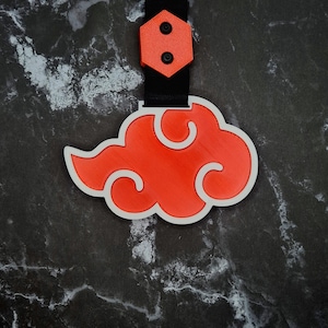 Puede incluir: Un llavero rojo en forma de nube con contorno blanco y correa negra. El llavero tiene un pequeño hexágono naranja con dos círculos negros. El diseño es una nube estilizada.