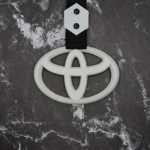 May include: A white Toyota logo keychain with a black strap and white backing. The Toyota emblem is a stylized oval design. The keychain is hanging against a dark, marbled background.