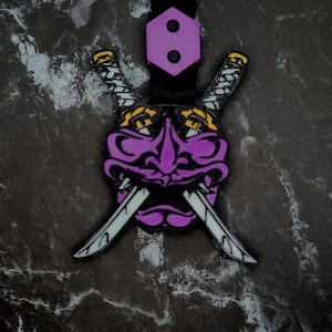 Könnte beinhalten: Eine lila-schwarze Oni-Maske mit zwei gekreuzten Schwertern. Die Maske hat gelbe Akzente und ist an einem schwarzen Riemen mit einem lila Sechseck befestigt. Das Design ist detailliert und stilisiert.