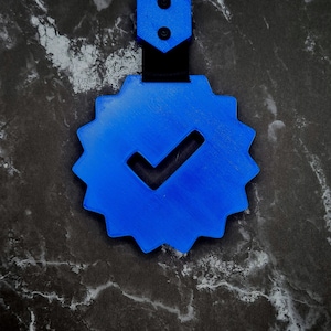 May include: A blue, star-shaped badge with a checkmark symbol. The badge is attached to a black strap and has two black rivets at the top. The background is a dark, marbled surface.