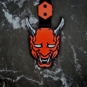 Op de afbeelding: Een 3D-geprinte oranje Oni-masker sleutelhanger met witte accenten en een zwarte band. Het masker heeft scherpe tanden en hoorns, met een zwarte zeshoekige bevestiging aan de bovenkant. De sleutelhanger staat tegen een donkere, getextureerde achtergrond.