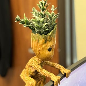 Puede incluir: Una pequeña figura de madera marrón de un personaje de la película Guardianes de la Galaxie. La figura está agarrada al borde de un monitor de computadora. La figura tiene una pequeña planta verde que crece de su cabeza.
