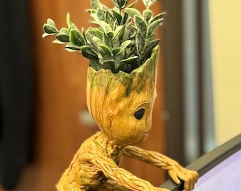 May include: A small, brown, wooden figurine of a character from the movie Guardians of the Galaxy. The figurine is holding onto the edge of a computer monitor. The figurine has a small green plant growing out of its head.