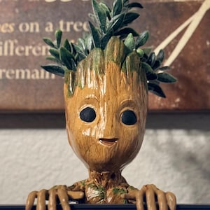May include: A brown and green ceramic planter shaped like the character Groot from the Guardians of the Galaxy movie franchise. The planter has a smiling face and is holding a small succulent plant.