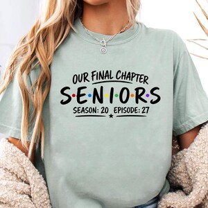 May include: A light sage green short-sleeve t-shirt featuring black text that reads Our Final Chapter Seniors Season 20 Episode 27. The word Seniors is written with colorful dots between each letter, and a small star is centered below the text.