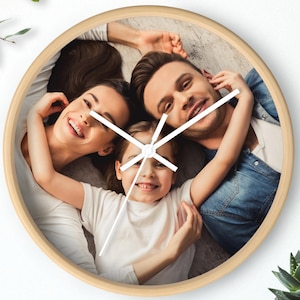May include: Round wall clock with a light wood frame. The clock face features a family portrait of a smiling family. White clock hands indicate the time. A small potted plant sits on a wooden surface.