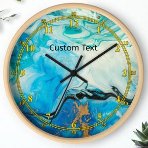 May include: Round wall clock with a light wood frame. The clock face features a blue and white abstract design with gold accents and black hands. The text "Custom Text" is printed on the clock face.