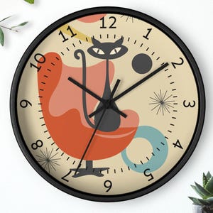 May include: A black-framed wall clock with a mid-century modern design. The clock face features a stylized black cat on an orange chair, with abstract shapes in red, blue, and yellow. The clock hands are black.