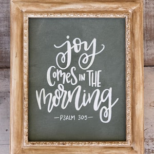May include: A framed canvas print with a green background and white lettering that reads "joy comes in the morning - PSALM 30:5 - ". The frame is a light brown wood with a decorative edge.