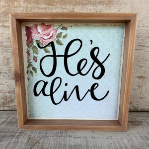 May include: Wooden framed sign with the words "He's alive" in black script. The background is a light blue with a floral design and a floral corner design with pink roses and green leaves.