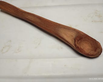 Op de afbeelding: Een handgemaakte houten lepel met een lange steel en een afgeronde kom. De lepel is gemaakt van roodbruin hout, dat de natuurlijke nerf en textuur laat zien. De lepel is ontworpen om te koken en te serveren.