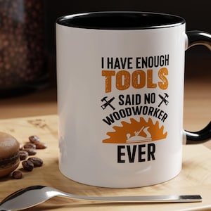 May include: White ceramic mug with a black rim and handle. The mug has a graphic that says "I have enough tools said no woodworker ever" with a saw blade and a plane.