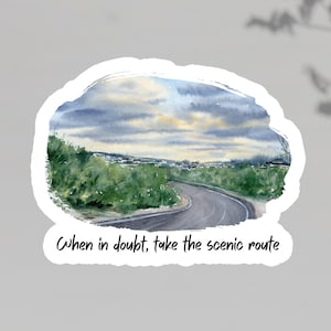 May include: A sticker with a watercolour painting of a road winding through a green landscape under a cloudy sky. The text "When in doubt, take the scenic route" is written below the image in black script.