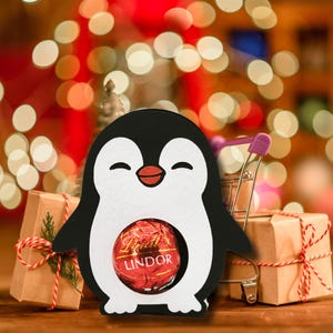 May include: A black and white penguin-shaped gift box holding a red Lindor chocolate ball. The penguin has a smiling face. Two wrapped gifts and a miniature shopping cart are in the background.