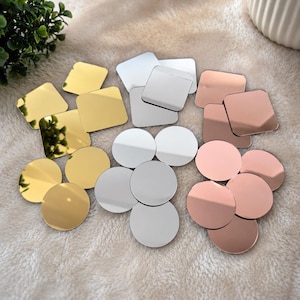 May include: A collection of small, reflective acrylic shapes in gold, silver, and rose gold finishes. The set includes both square and circular pieces with smooth, polished surfaces that create clear reflections of the surrounding environment.