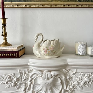 May include: A cream-coloured ceramic swan planter featuring a textured feather design and a hand-painted floral motif on its side. The swan has a curved neck and an open top, with delicate pink and blue flowers detailed on the central body area.