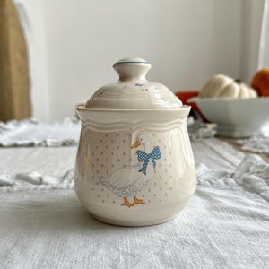 Könnte beinhalten: Eine cremefarbene Zuckerdose aus Keramik mit passendem Deckel, verziert mit einem Muster aus kleinen braunen Blumen und einer zentralen Abbildung einer weißen Gans mit einer blauen gepunkteten Schleife, orangefarbenem Schnabel und Füßen.