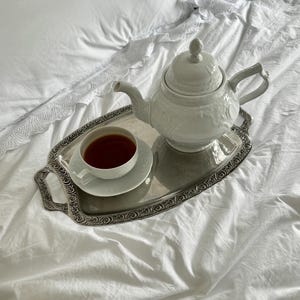 May include: A white porcelain teapot and teacup with dark tea sit on a silver tray. The tray is placed on a white bedspread. The teapot has a decorative floral pattern and a lid with a knob.