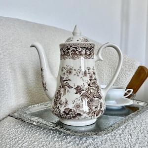 Puede incluir: Una tetera de cerámica color crema con un patrón marrón de flores, follaje y un pájaro. La tetera tiene una tapa a juego con un remate, un asa curva y un pico largo, todo ello sobre una bandeja rectangular de metal de tono plateado.