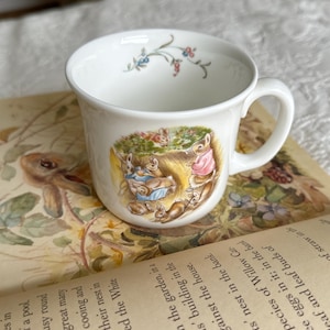 May include: White ceramic mug with a handle, featuring a colorful illustration of rabbits in a garden scene. Delicate floral sprigs adorn the rim. The mug is set on an open book with text visible.