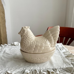 Könnte beinhalten: Ein cremefarbener Keramikbehälter in Form eines Huhns. Die obere Hälfte ist ein detailliertes Huhn, und die untere Hälfte ist eine korbähnliche Schüssel. Der Behälter steht auf einer hellgrauen Rüschen-Tischdecke. Der Hintergrund umfasst einen Tisch und einen Stuhl.