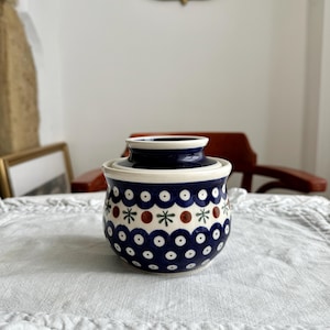 Puede incluir: Una mantequillera redonda de cerámica con un esmalte azul profundo y un patrón repetitivo de círculos blancos, pequeños motivos en forma de estrella verde y puntos marrones. El recipiente tiene una base ancha y una tapa a juego con borde acampanado, mostrando una estética pintada a mano con un acabado brillante.