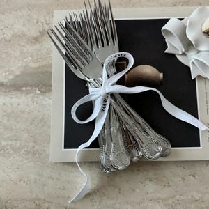 May include: A collection of silver forks, tied together with a white ribbon bearing the word "LOEWE". The forks have decorative handles and are presented on a black and white card.
