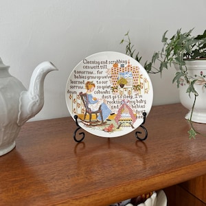 Könnte beinhalten: Ein dekorativer Teller mit Kreuzstichmuster, der eine Frau in einem Schaukelstuhl und Text zeigt. Der Teller wird auf einem schwarzen Metallständer ausgestellt. Der Text auf dem Teller lautet "Cleaning and scrubbing can wait till tomorrow..."