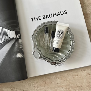 May include: A silver-toned, scalloped-edge metal dish containing two small clear glass perfume vials and one cream-colored tube of Diptyque Voile Satin Oil for Body and Hair, 10 ml, 0.34 fl. oz., resting on a book titled The Bauhaus by Magdalena Droste.