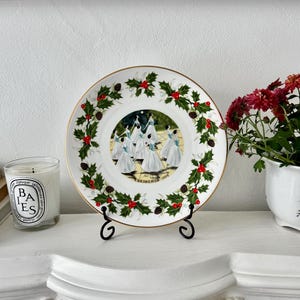May include: A decorative plate with a gold rim, featuring a central image of dancers in white dresses. The plate is adorned with holly, red berries, and pine cones. The plate is displayed on a black metal stand.