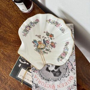 May include: A white, shell-shaped decorative dish with a floral and bird design, accented with gold trim. A gold necklace rests in the dish, which is placed on a book with the text "Queen Victoria". A bottle of perfume sits nearby.