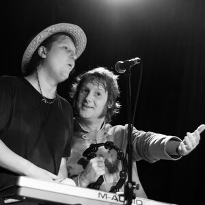 May include: Two men, one wearing a straw hat, are playing music together. The man in the hat is playing a keyboard, while the other man is playing a tambourine. They are both smiling and enjoying themselves.