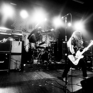 May include: Black and white photo of a rock band performing on stage. The band members are playing electric guitars and drums. The stage is lit with spotlights.