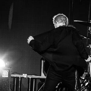 May include: A black and white photograph of a person on stage, seen from the back, pulling open a black jacket. Stage lighting illuminates the scene, with a drum set visible in the background.