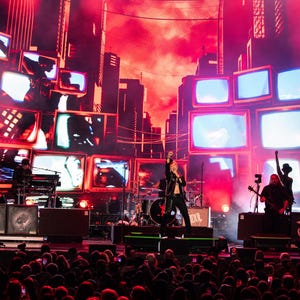 May include: A live concert stage with a backdrop of vintage televisions and a cityscape silhouette. The stage is lit with red and white lights, and a band is performing in front of a crowd. The band members are playing instruments and singing.