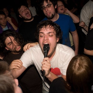 May include: A group of people are gathered together, some are holding microphones and singing. The person in the center is wearing a white t-shirt and holding a microphone. The person on the right is wearing a black t-shirt with the text "COBB" printed on it.