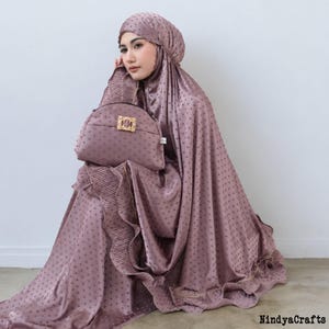 以下が含まれることがあります： 水玉模様の、頭巾とハンドバッグがセットになったモーヴ色の祈祷服。 プリーツとフリルが施され、ゴールドのトリムが施されています。 ハンドバッグには装飾的な留め金が付いています。「NindyaCrafts」の文字が見えます。