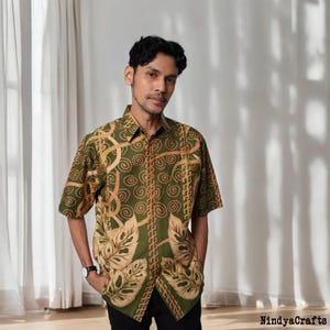 Puede incluir: Camisa de manga corta con botones, con fondo verde y estampado de hojas y espirales color beige. La camisa tiene cuello clásico y se lleva abierta. El texto "NindyaCrafts" es visible en la esquina inferior derecha.