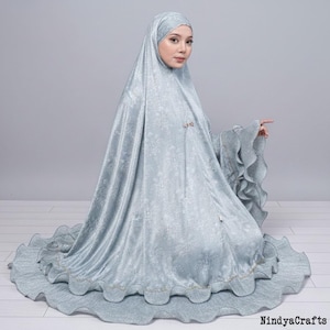 May include: A pale blue prayer garment featuring a floral design. The set includes a head covering and a long skirt with a ruffled hem. The sleeves have a pleated design with ruffled edges. The garment is made of a soft, silky material.