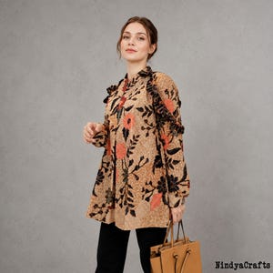 May include: A long-sleeved blouse featuring a tan base with a black and orange floral batik pattern, a button-down front, and ruffled shoulder details. The garment is paired with black trousers and a tan leather handbag. Text reads NindyaCrafts.