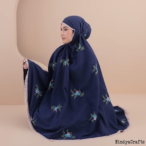 May include: A dark blue prayer garment featuring a matching head covering and a full-length silhouette. The fabric is adorned with small, scattered floral embroidery in shades of blue, green, and pink, finished with a light beige scalloped lace trim.
