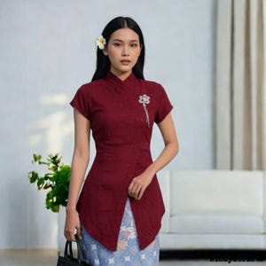 Op de afbeelding: Bordeauxrode top met korte mouwen, een mandarijnkraag en een decoratieve broche. De top heeft een split aan de voorkant en wordt gecombineerd met een rok met patroon. Een witte bloem siert het haar.