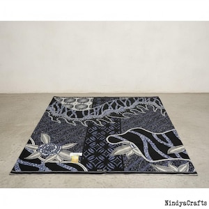 Op de afbeelding: Een rechthoekig batiktextiel met een donkerblauw en zwart bloemen- en geometrisch patroon. De stof heeft aan de ene kant een groot bloemendesign en aan de andere kant een draakachtig motief. Het textiel wordt gepresenteerd op een grijze ondergrond.
