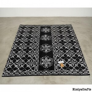 May include: Black and white patterned textile with a central panel featuring snowflake-like designs. The textile has a repeating geometric pattern and is laid out on a concrete surface. The text "AYTEX 003" is visible.