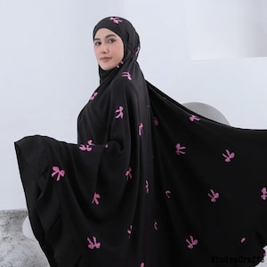 May include: Black prayer garment with pink bow accents. The garment includes a head covering and a flowing, full-length design. The bows are evenly spaced across the fabric. The garment is worn by a person.