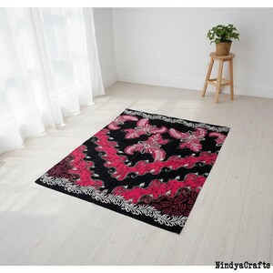 Op de afbeelding: Een rechthoekig zwart en rood patroon textiel vloerkleed met bloemmotieven. Het vloerkleed heeft een witte rand met een bloemmotief. Het vloerkleed ligt op een lichte houten vloer. Een kleine houten kruk met een plant staat op de achtergrond.