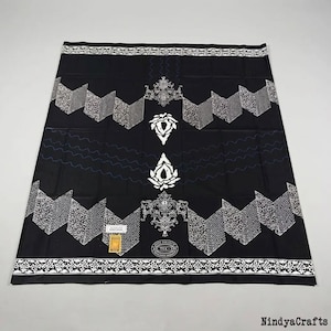 Op de afbeelding: Zwarte sarong met witte geometrische en bloemmotieven. De stof heeft een herhalend zigzagontwerp langs de randen en een centraal bloemmotief. De sarong heeft een decoratieve rand en een label.