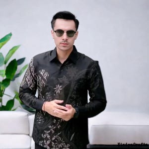 May include: A black button-down shirt with a batik pattern. The shirt features a floral and geometric design in shades of black and tan. The long-sleeved shirt is worn with sunglasses.